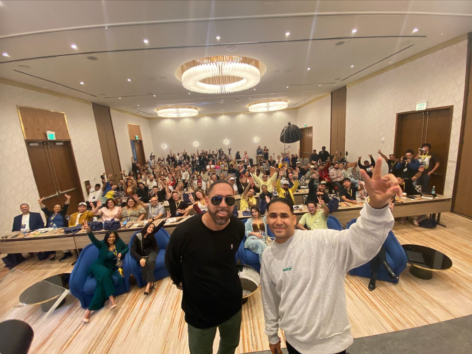 A packed conference room of attendees cheering and waving behind two smiling hosts at The Latin Music Hub Conference